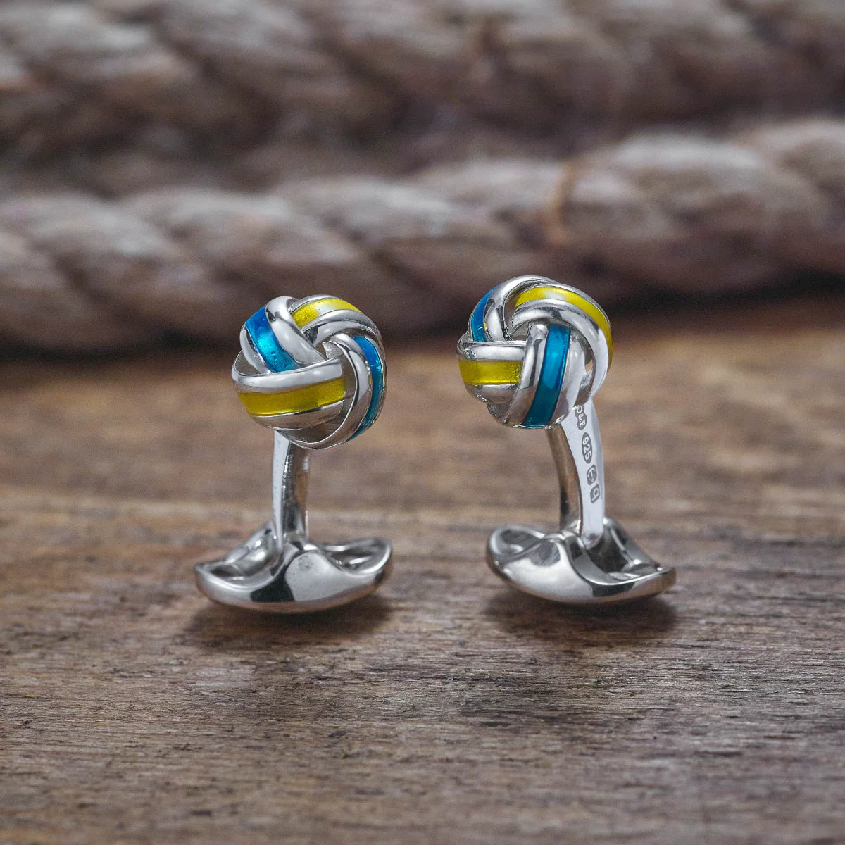 Silver knot cufflinks with blue and yellow accents on a wooden surface Deakin & Francis - EB London
