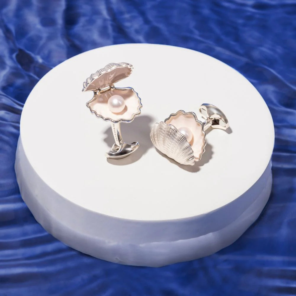 Shell-shaped cufflinks with a pearl on a white stand against a blue water background