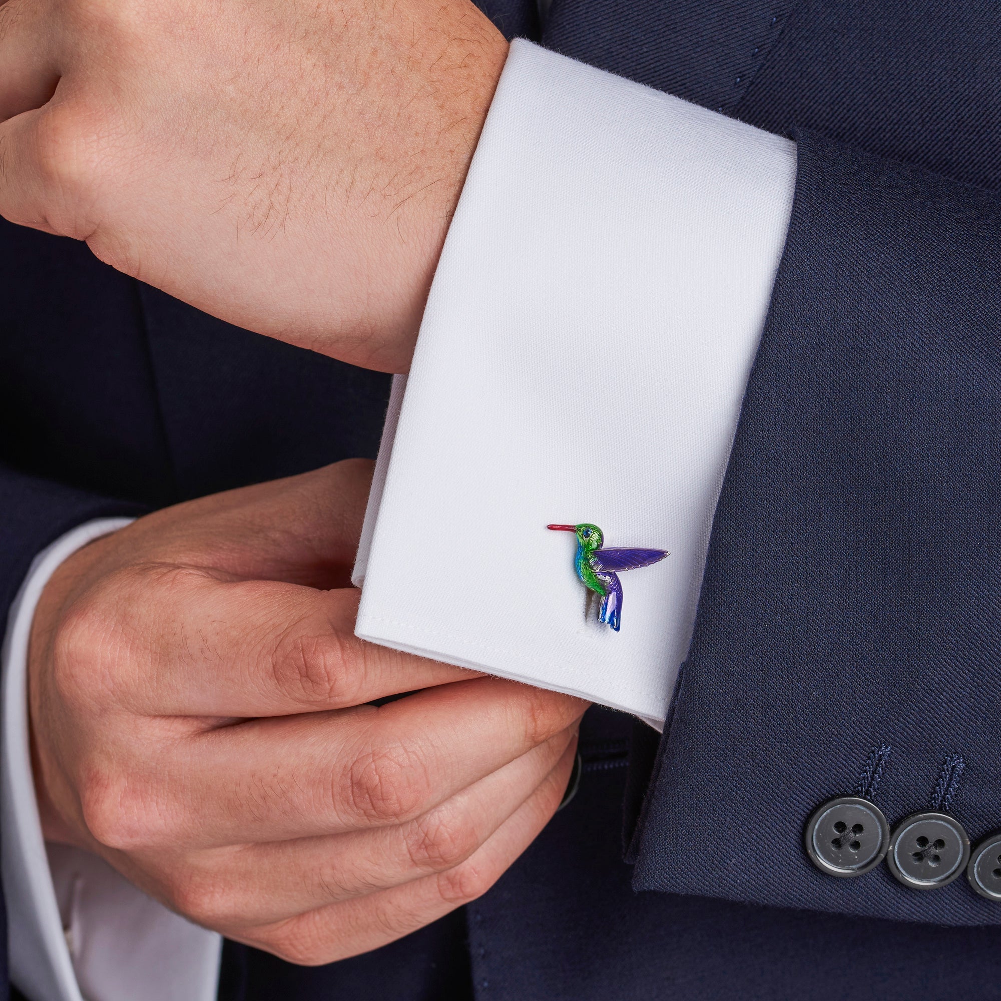 Close-up of a person adjusting a cufflink on a white shirt sleeve with a navy suit. Hummingbird - EB London